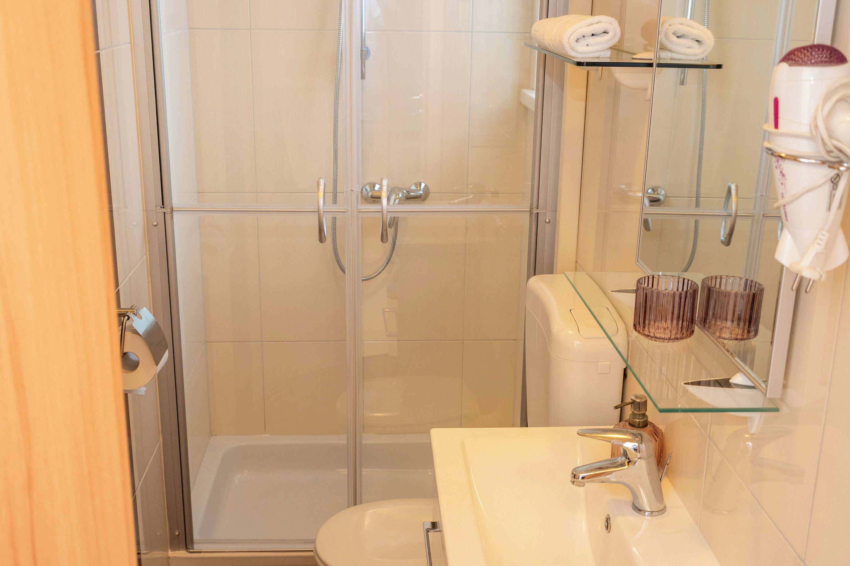 A bathroom with a glass shower enclosure, a toilet, a sink with a glass shelf, and a mirror.