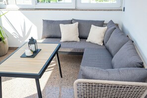 Ein sonnendurchfluteter Patio-Bereich mit einem grauen Sofa, einem kleinen Tisch und einem Korbstuhl.