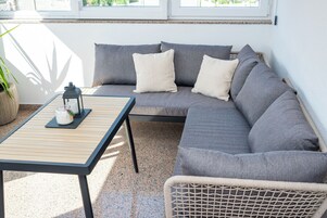 Un patio iluminado por el sol con un sofá gris, una mesita y una silla de mimbre.