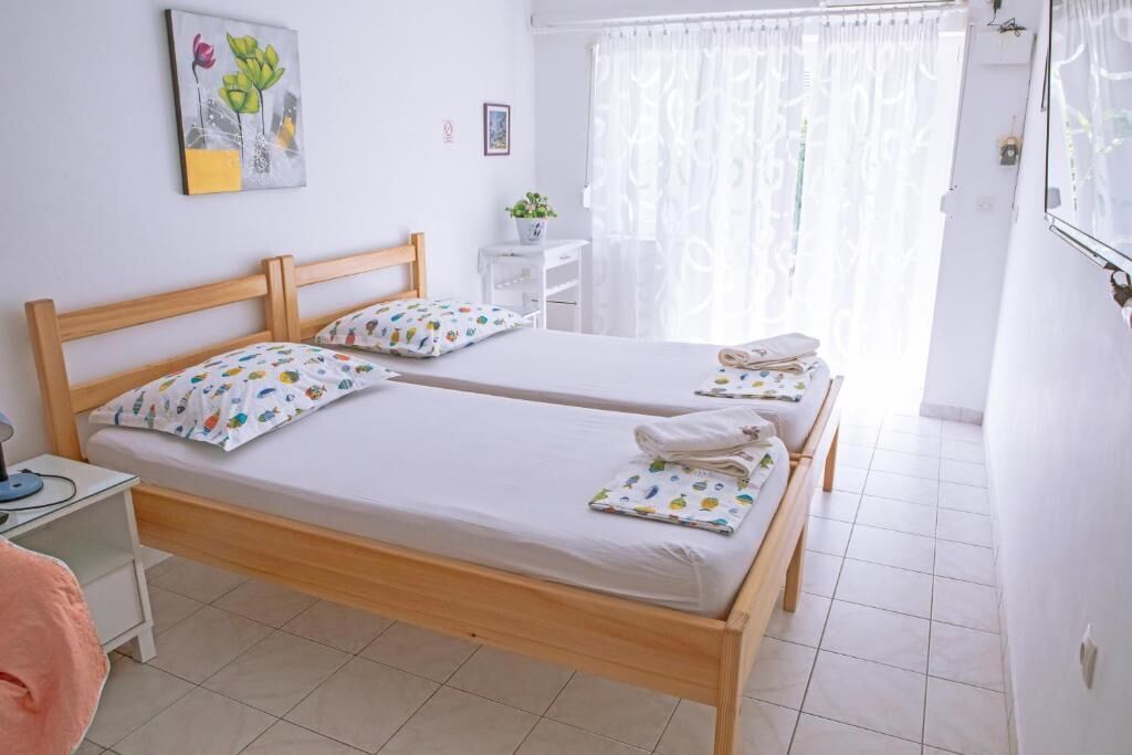 Una cama doble con cabecera de madera, una mesita de noche, una ventana con cortinas traslúcidas y un cuadro colgado en la pared.