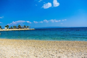 A beach with pebbles, clear blue water, and a structure with palm trees. - Apartments Studio Spalato (Split)
