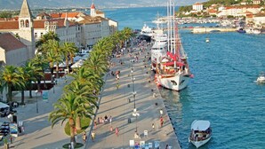 A waterfront promenade with a large ship docked, palm trees lining the path, and historic buildings in the background. - Apartments Kate (Trogir)