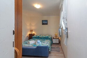 Ein schmaler Raum mit einem Bett, einem Nachttisch, einer Lampe und einem Bild an der Wand.