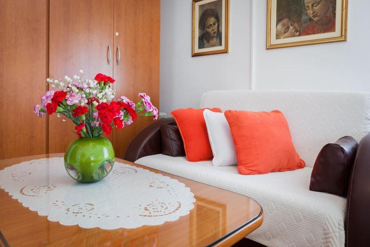 Ein Zimmer mit einem Holzschrank, einem weißen Sofa mit orangefarbenen und weißen Kissen, einer grünen Vase mit Blumen und zwei gerahmten Bildern an der Wand.