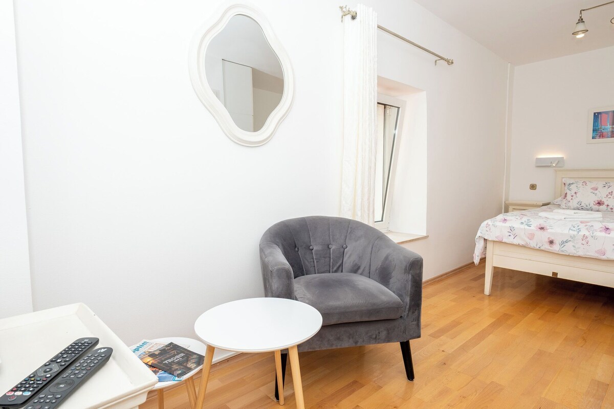 Ein Zimmer mit grauem Sessel, einem kleinen weißen Tisch, einem Bett mit Blumenmuster auf der Bettwäsche, einem weißen Spiegel und einer Tür mit Vorhängen.