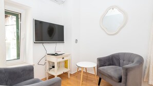 Ein Wohnzimmer mit Flachbildfernseher, zwei grauen Sesseln, einem kleinen weißen Tisch und einem runden Spiegel an der Wand.