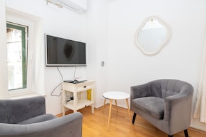 Ein Wohnzimmer mit Flachbildfernseher, zwei grauen Sesseln, einem kleinen weißen Tisch und einem runden Spiegel an der Wand.
