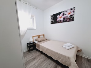A bedroom with a bed, a nightstand, a window with curtains, and a wall-mounted picture of flowers. - Apartment Sia (Crikvenica)