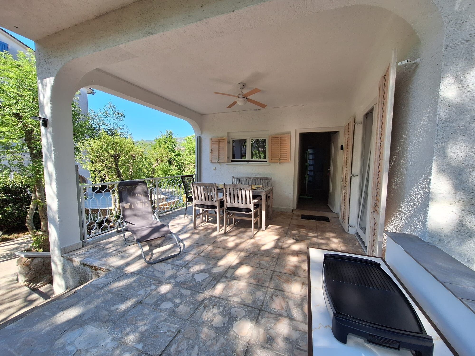 Une terrasse extérieure couverte, dotée d’une table et de chaises, d’un ventilateur de plafond et offrant une vue sur la verdure.