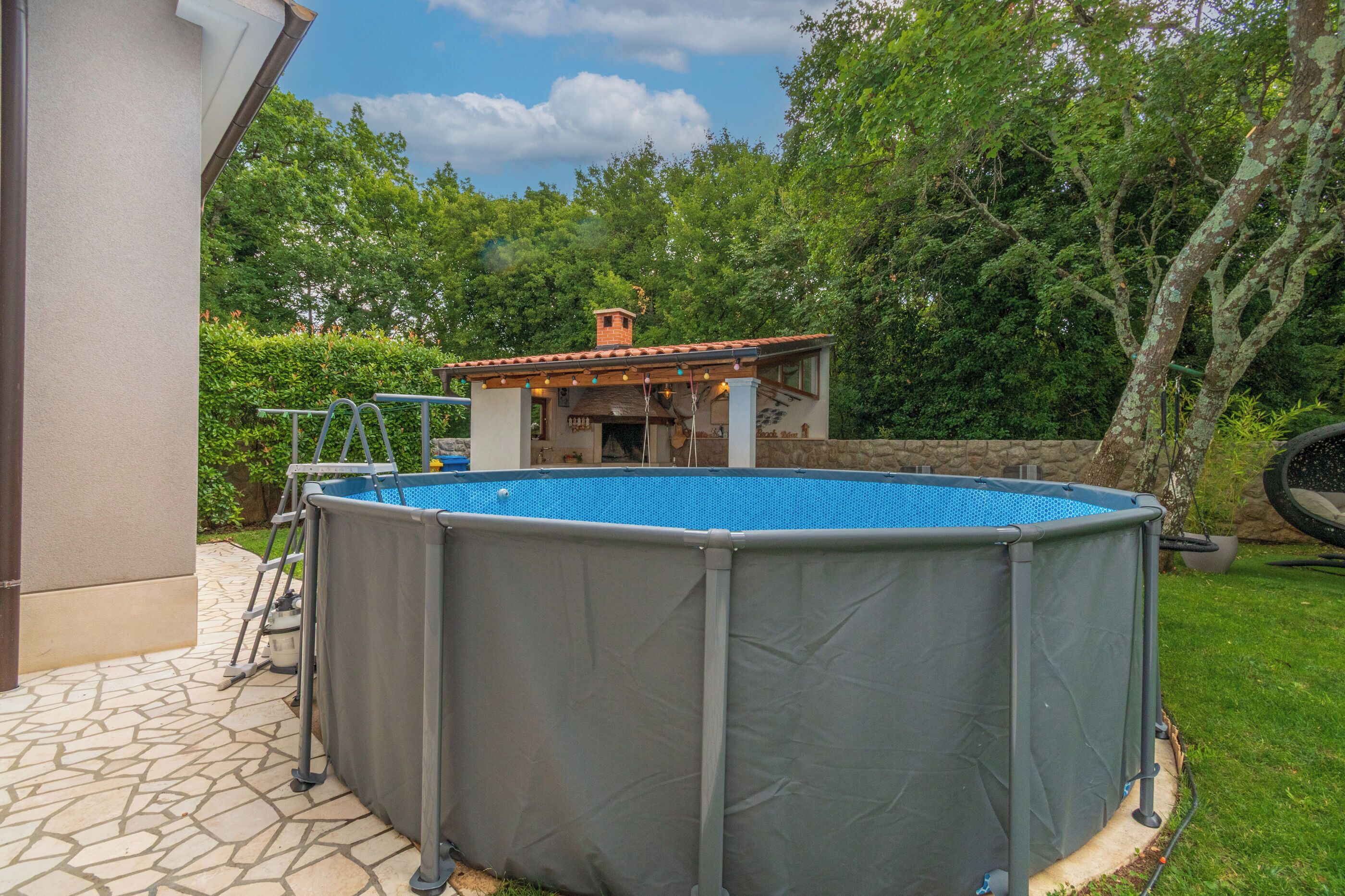 Una piscina fuori terra con una scaletta, una casetta con tetto in tegole e un vialetto in pietra.