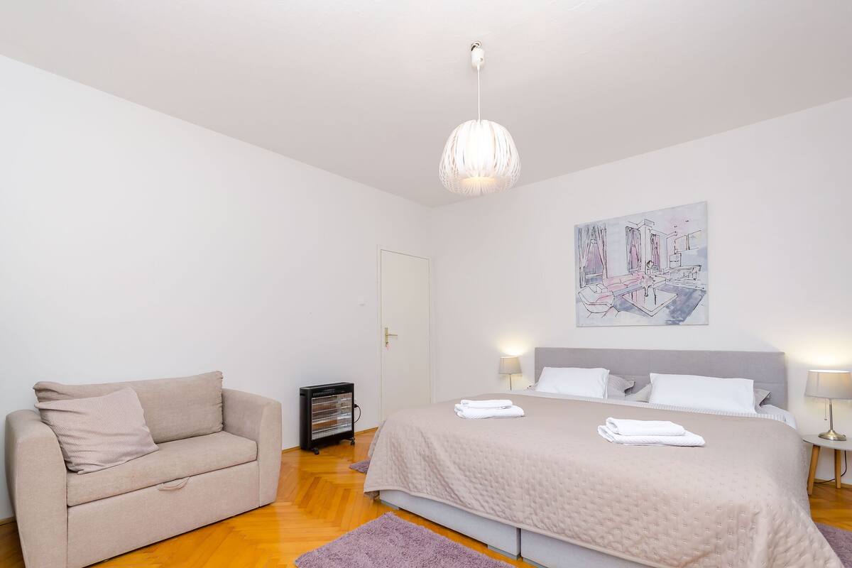 A bedroom with a bed, a sofa, a heater, a painting, and a lamp.