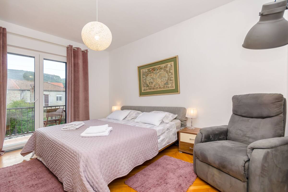 A bedroom with a bed, a chair, a nightstand, a wall-mounted lamp, and a window with curtains.