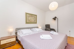 Ein ordentlich bezogenes Bett mit weißen Leinen, eine Nachttischlampe, ein Sessel und ein an der Wand hängendes Kunstwerk.