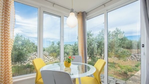 Ein Zimmer mit Glastisch, gelben Stühlen, einem weißen Stuhl, einer Hängeleuchte und Blick ins Grüne.