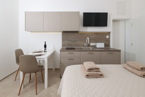 Ein kleines Hotelzimmer mit Bett, Schreibtisch, Stuhl, einer kleinen Küchenzeile und einem Fernseher.