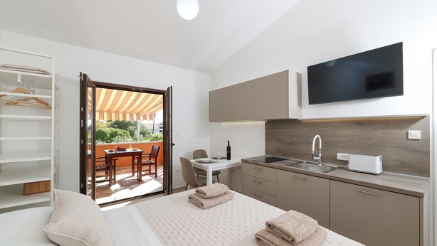 A modern hotel room with a bed, a TV, a kitchen area, and a balcony with a table and chairs. - Apartments Andi (Sukosan)