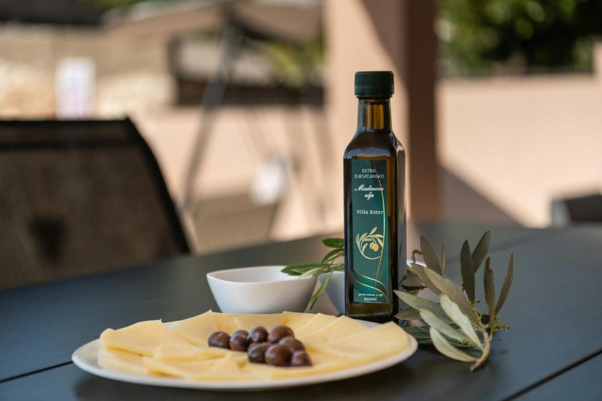 A bottle of extra dijevanski oil, a plate with cheese and olives, and a bowl on a table.