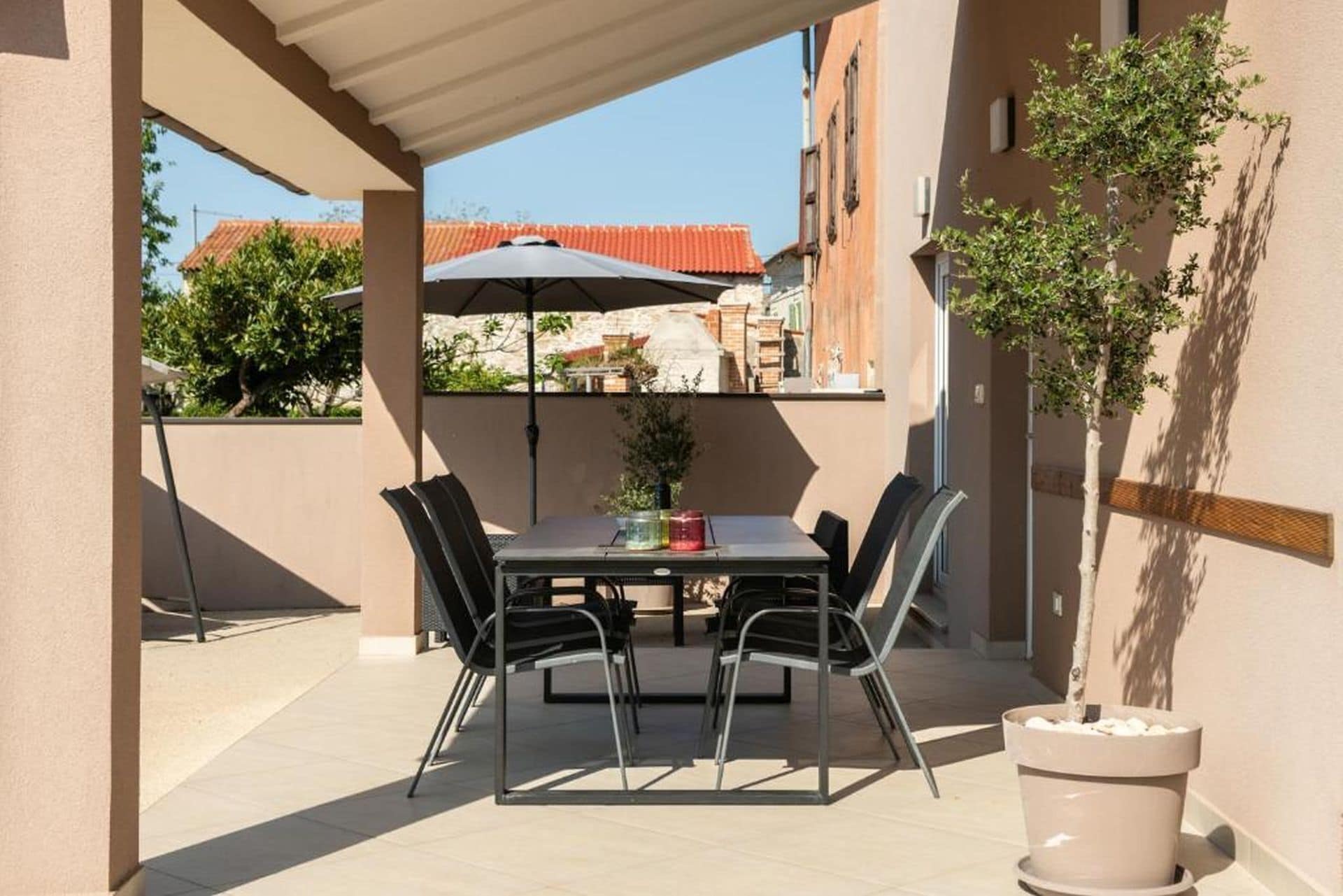 Un espace extérieur abrité, aménagé pour les repas, avec une table et des chaises, un pot de fleurs et une vue sur les bâtiments en arrière-plan.