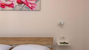 A bedroom with a wooden headboard, white pillows, a bedside shelf with a plant, a power outlet, and a painting of flowers. - House Ester (Liznjan)