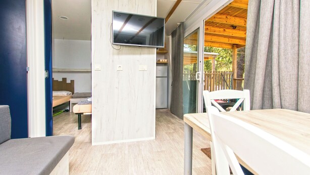 A room with a bed, a TV mounted on a wall, a table, and a chair. - Mobile Home Fisherman (Pasman)