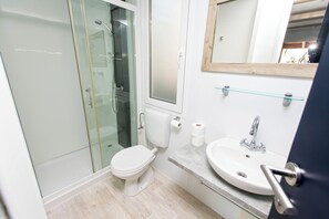 Ein Badezimmer mit Duschkabine aus Glas, Toilette, Waschbecken und einem Spiegel darüber sowie einem Glasregal.
