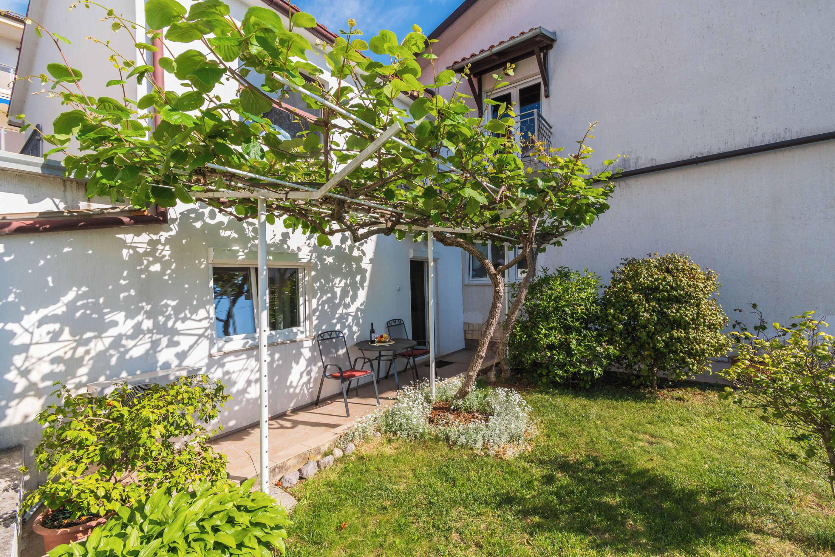 Um jardim com pérgola, vasos de plantas e um alpendre com cadeiras.