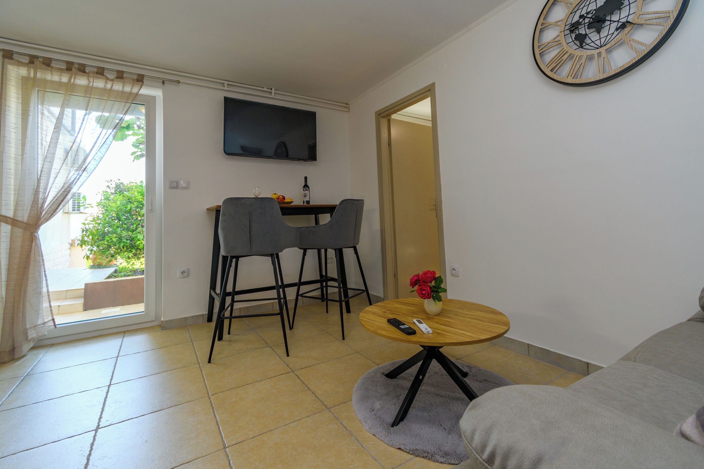 Una sala de estar con un sofá, una mesa de comedor con sillas, un televisor montado en la pared y un reloj.