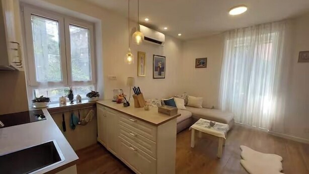 Ett modernt kök med en central köksö, en matplats med soffa och soffbord samt ett vardagsrum med ett stort fönster.