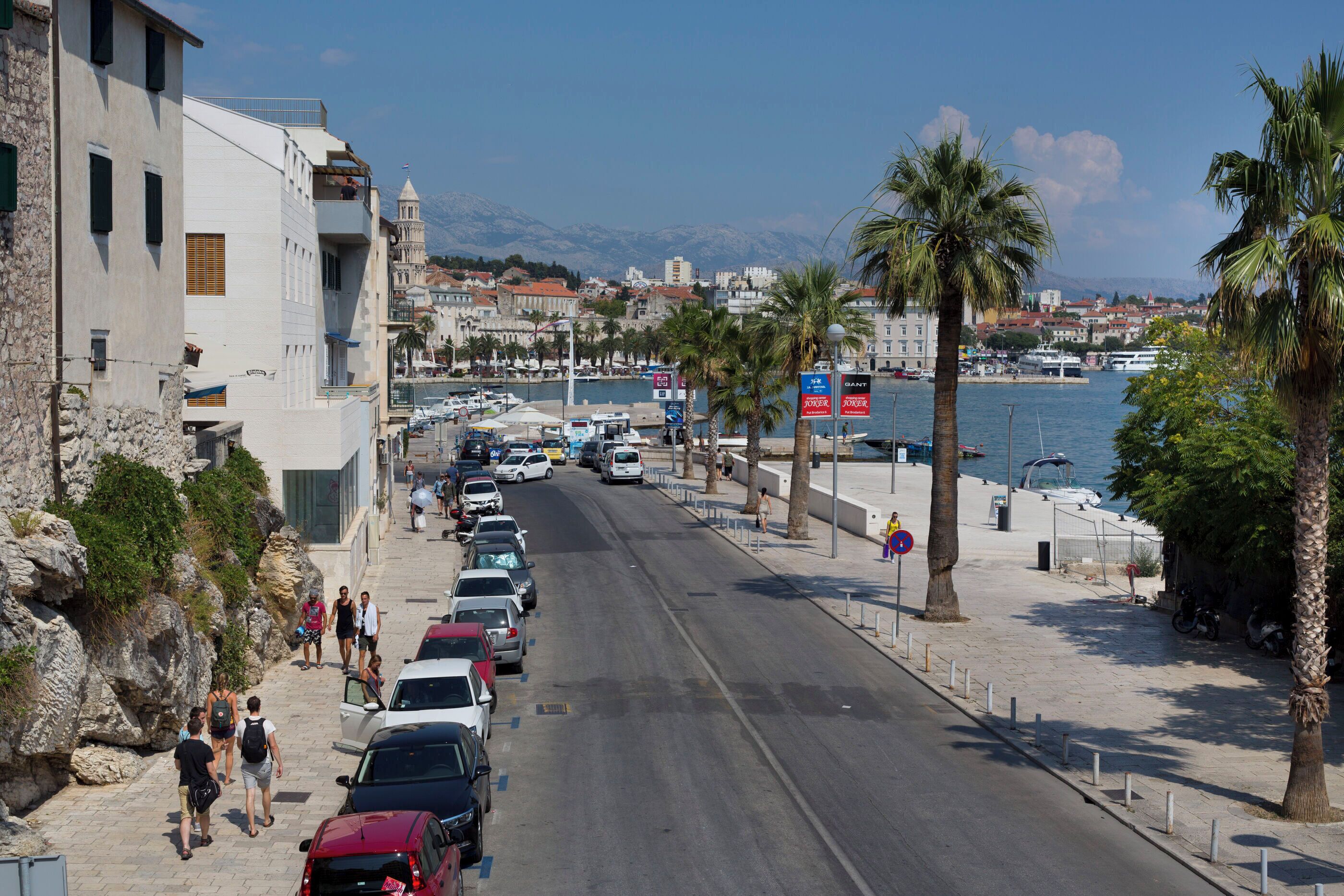 A coastal street with parked cars, a marina, and buildings along the waterfront.