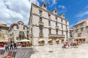 Ein historischer Platz mit einem markanten Gebäude, das Rundbogenfenster und Balkone aufweist, umgeben von Cafés und Terrassen.