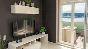 En moderne stue med et fladskærms-TV, et hvidt konsolbord og en balkon med udsigt over en bro og bakker.