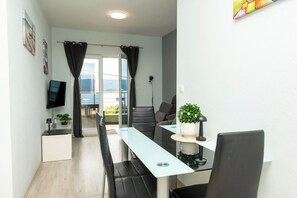 Ein modernes Wohnzimmer mit Glastisch, schwarzen Stühlen, einem Flachbildfernseher, einer Zimmerpflanze und Blick ins Freie.