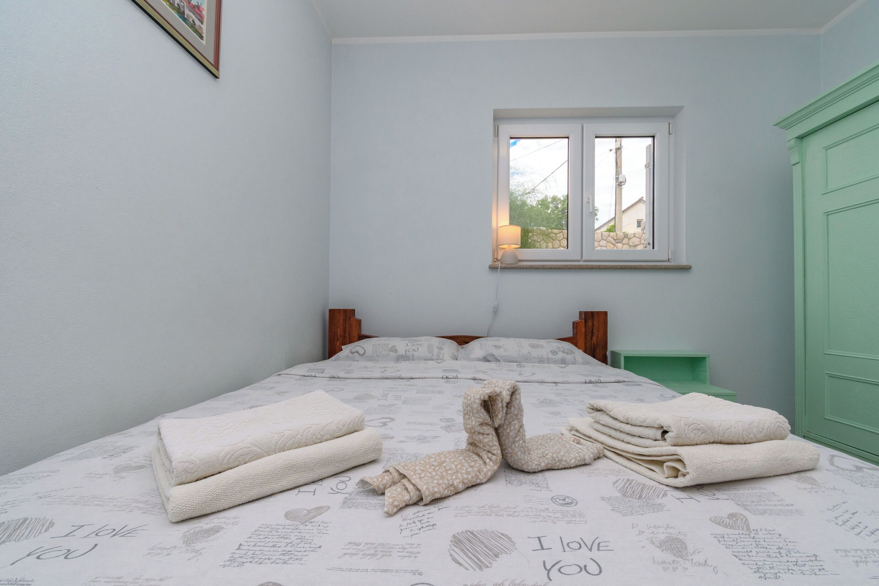 A neatly made bed with folded towels, a window with a view, and a green door.