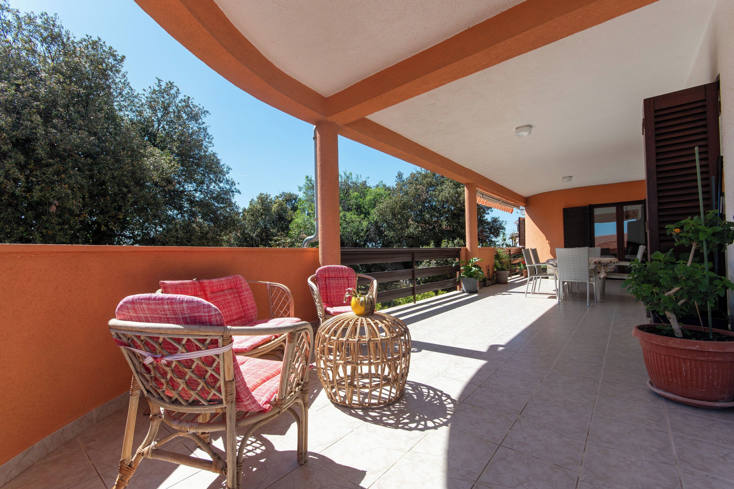 A covered outdoor patio with wicker furniture, potted plants, and a view of trees.