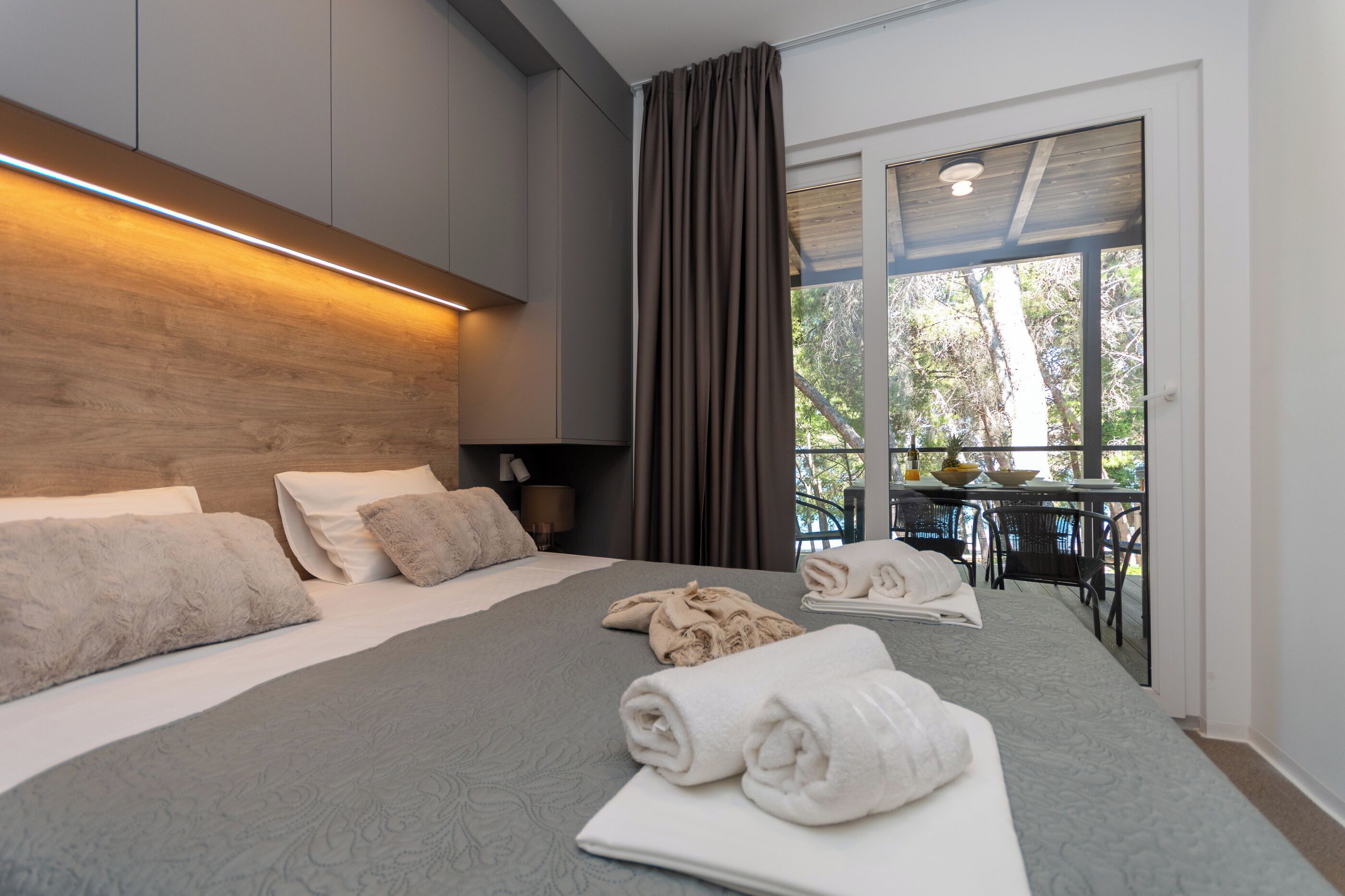 A modern hotel room with a large bed, a balcony with a table and chairs, and a view of trees.