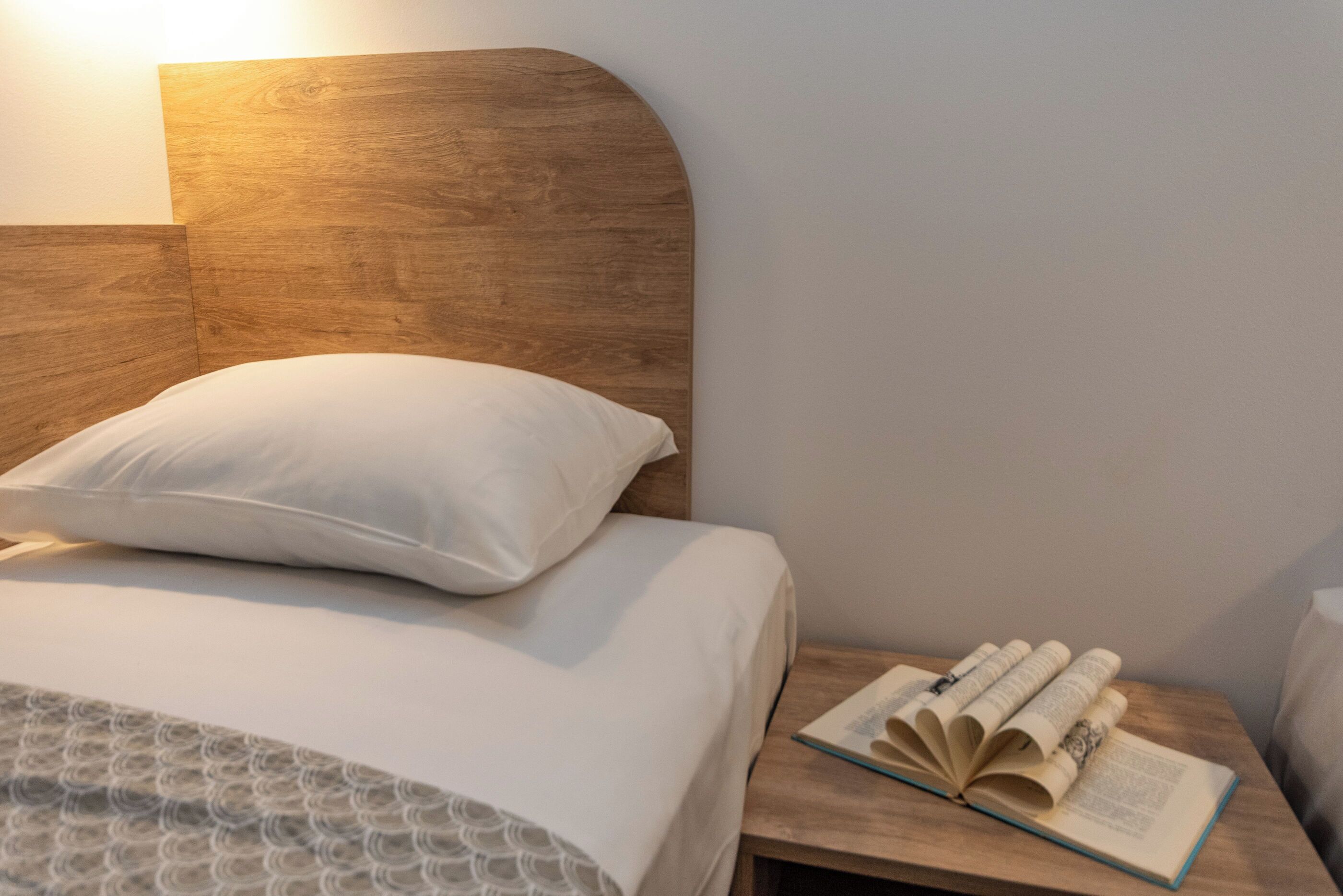 Un lit bien fait, avec un oreiller, une tête de lit en bois et une table de chevet sur laquelle repose un livre ouvert.