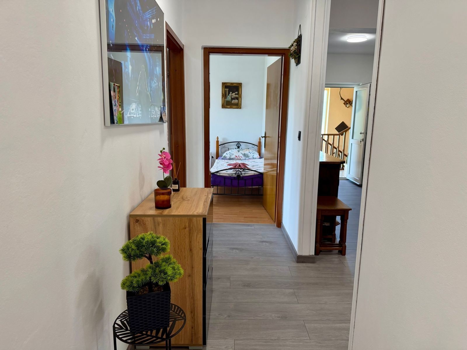Un couloir étroit mène à une chambre à coucher qui contient un lit, un bureau en bois, une plante en pot et un tableau encadré accroché au mur.
