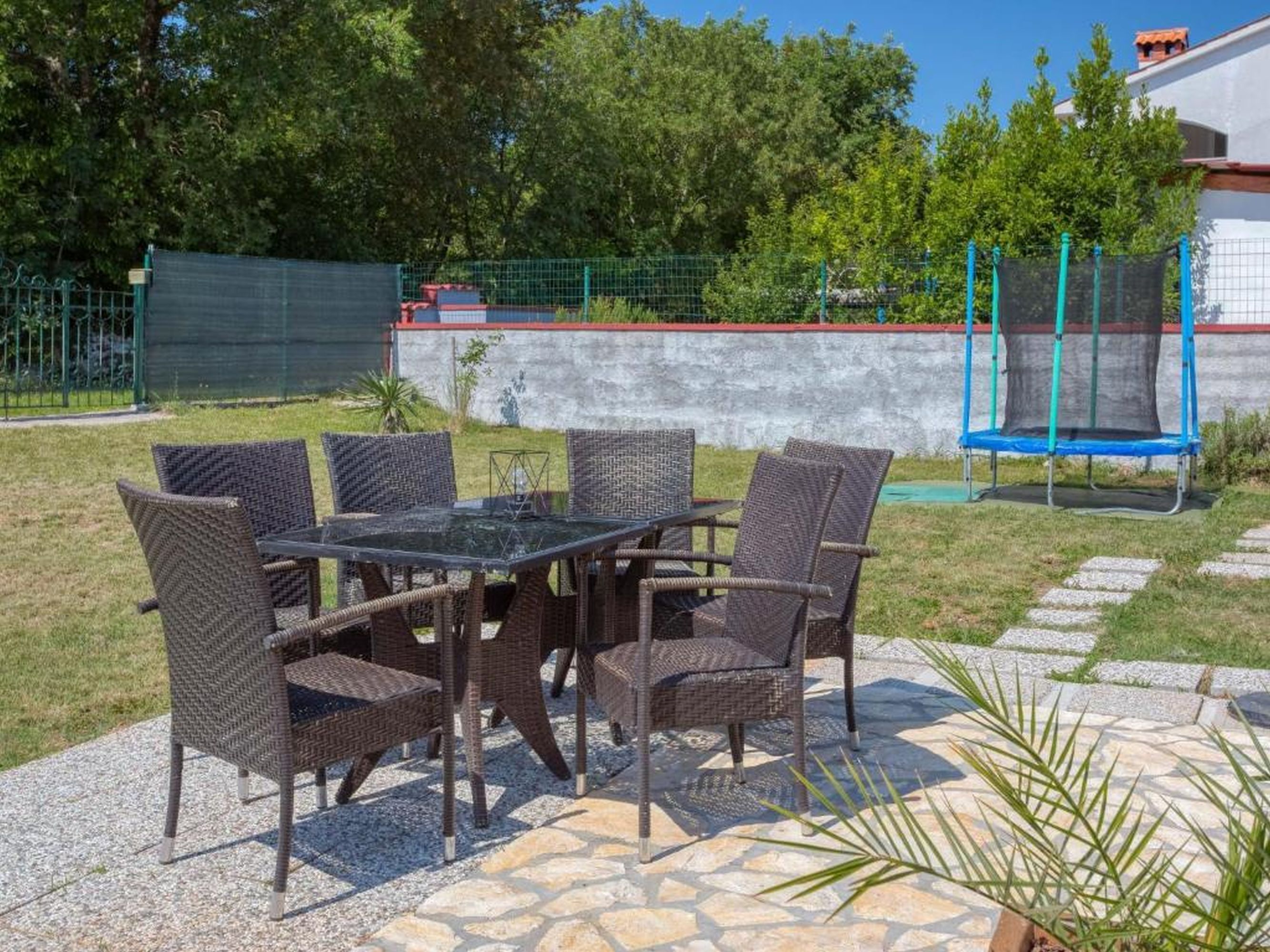 Un patio al aire libre con mobiliario de mimbre y una mesa de vidrio, rodeado por un sendero de piedra y follaje.