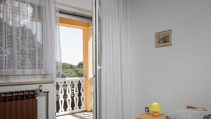 A bedroom with a bed, a bedside table with a lamp, a radiator, and a window with curtains. - House House Lola (Rasa)