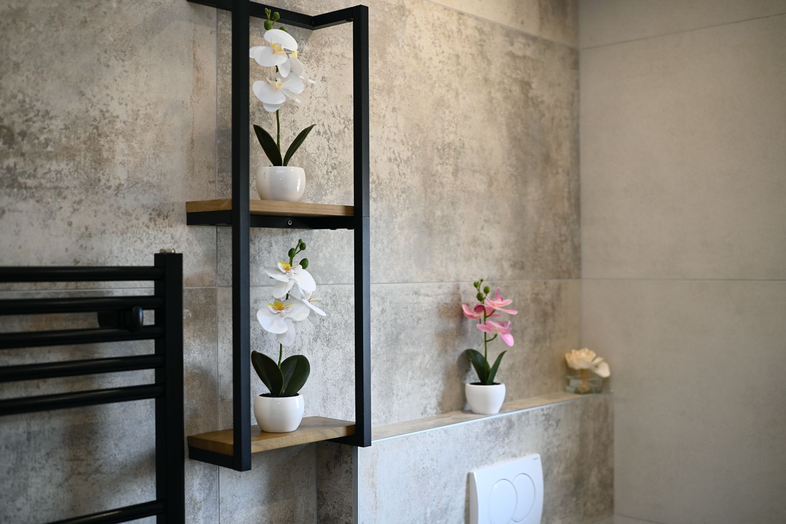 Ein Badezimmer mit einem schwarzen Metalletagenregal, das weiße und rosafarbene Orchideen, eine weiße Heizung und einen Holzstuhl beherbergt.
