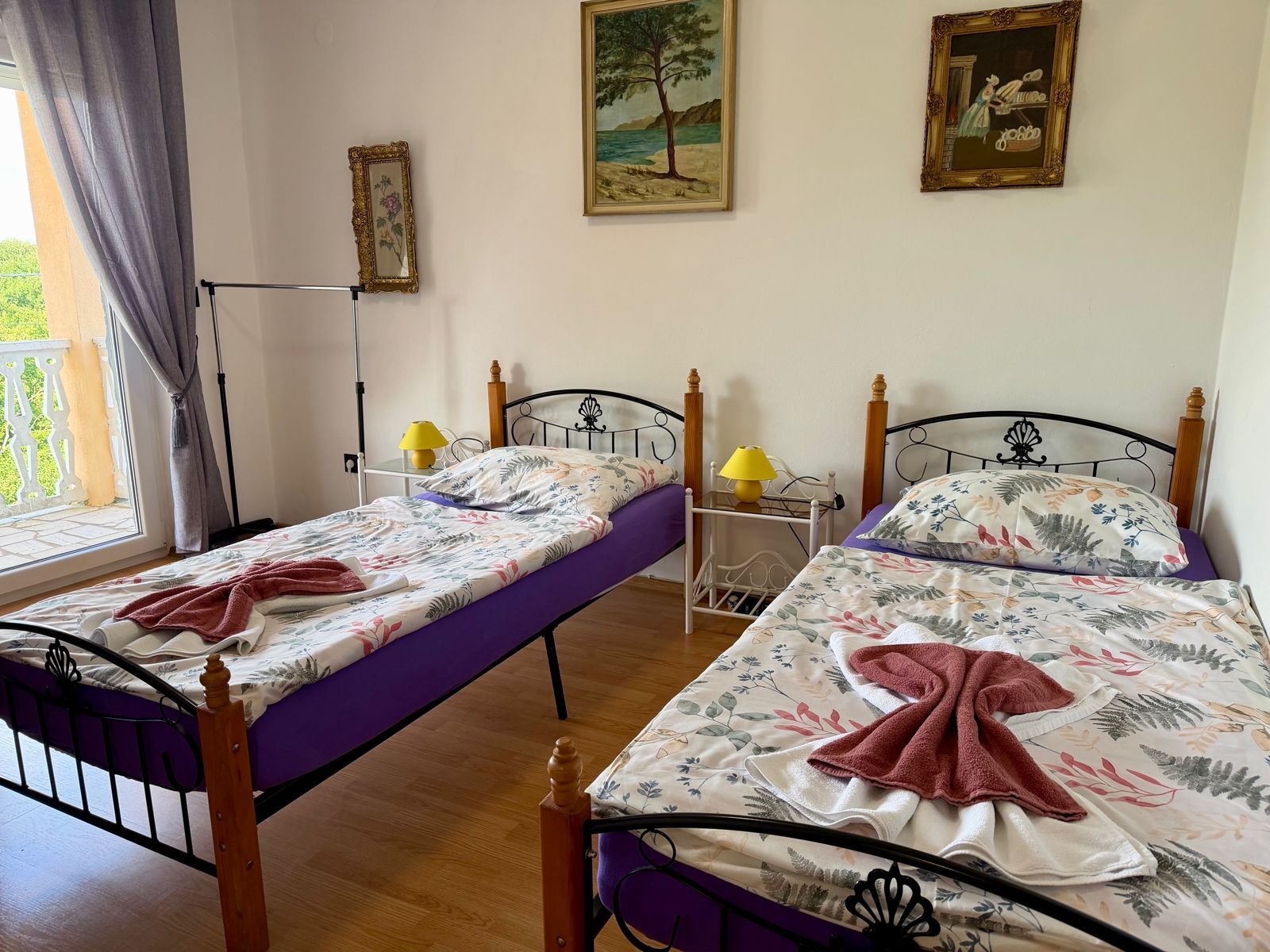 A room with two beds, each with a wooden headboard, a bedside table with a lamp, and a window with curtains.