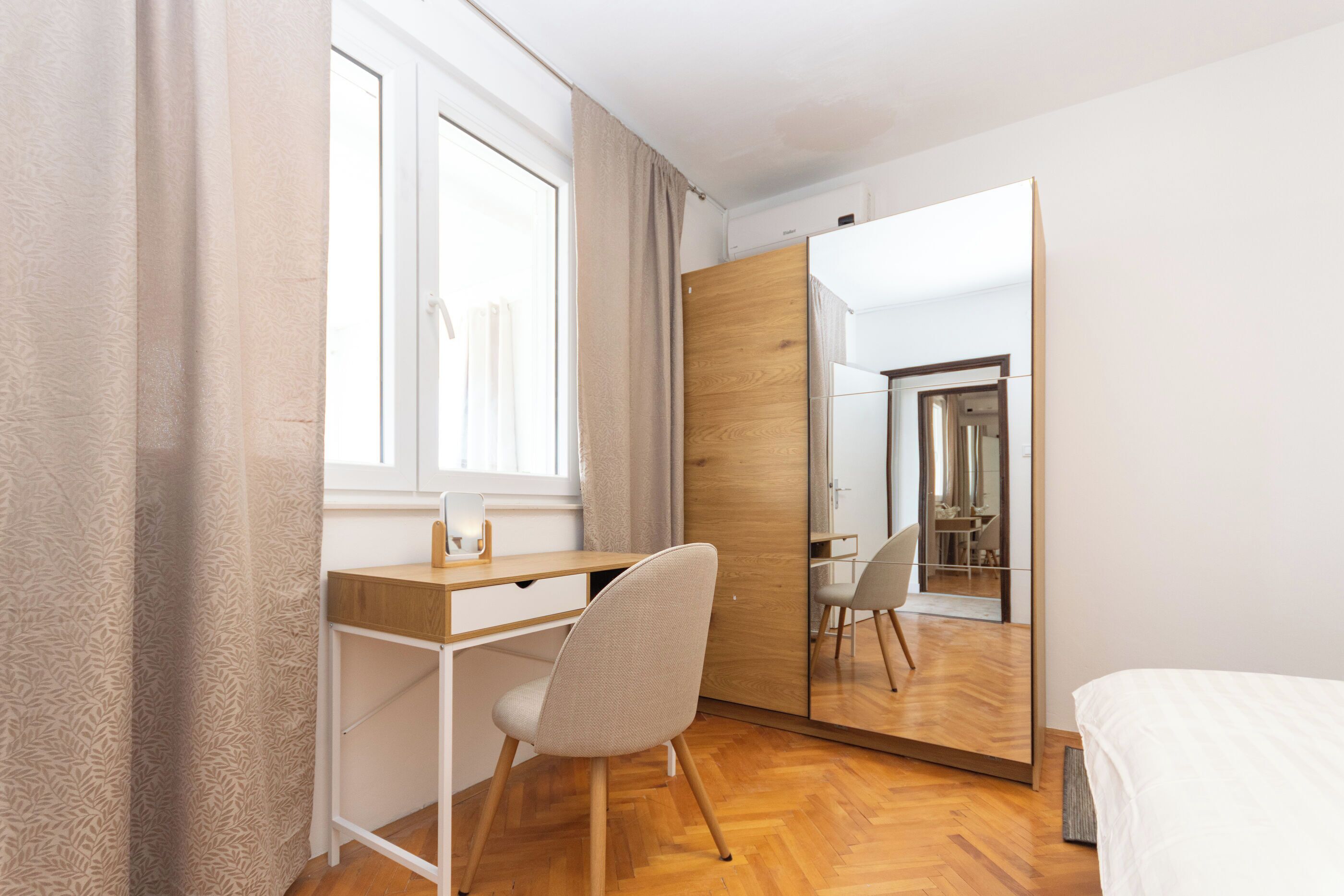 A bedroom with a wooden desk, a chair, a bed, a large mirror, and a wardrobe.