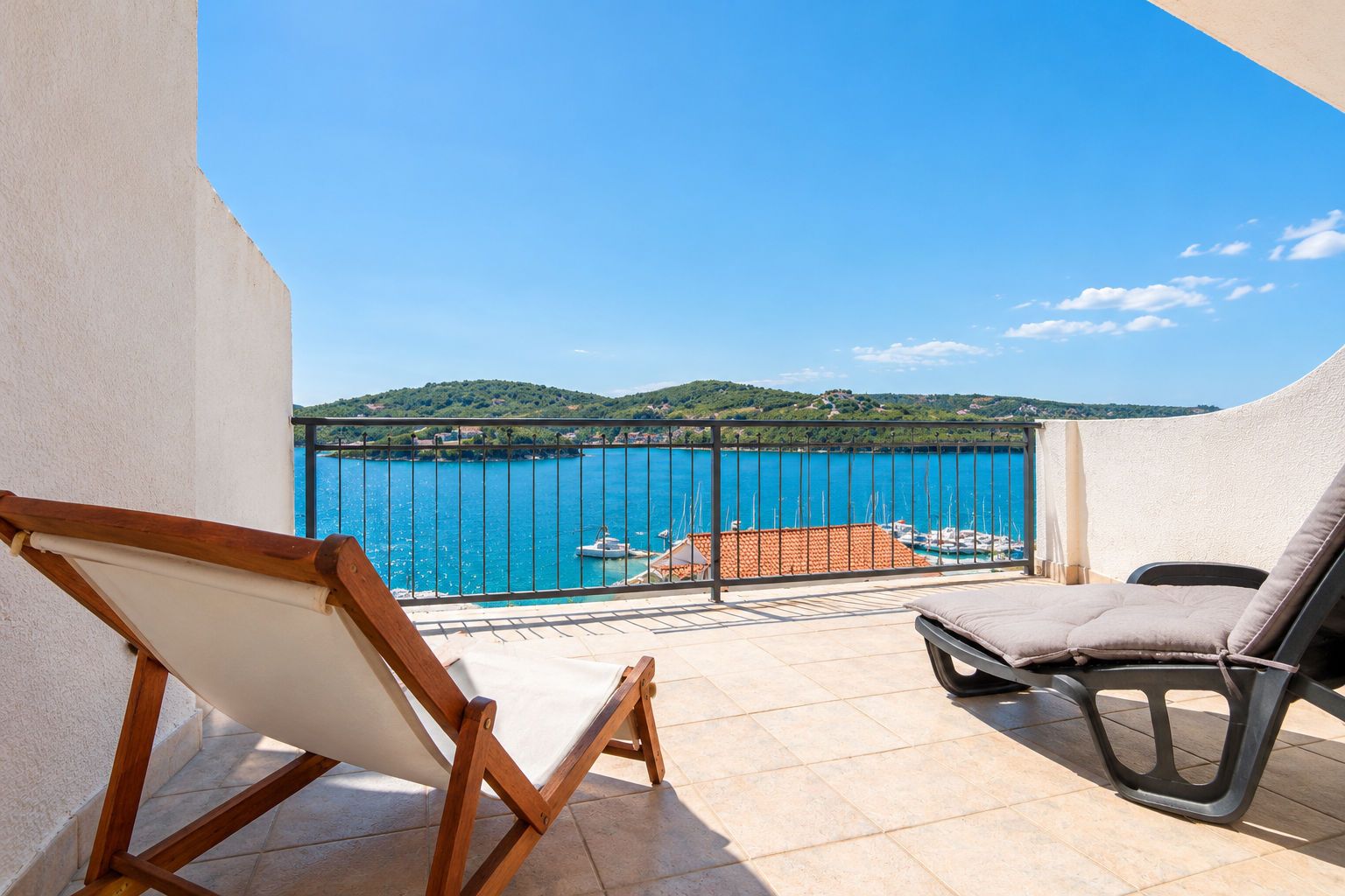 Un balcon avec vue sur une mer bleue et un paysage vallonné. On y trouve deux chaises et un chevalet en bois.