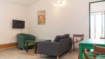 A living room with a sofa, a television mounted on the wall, a painting, and a dining area with a table and chairs.