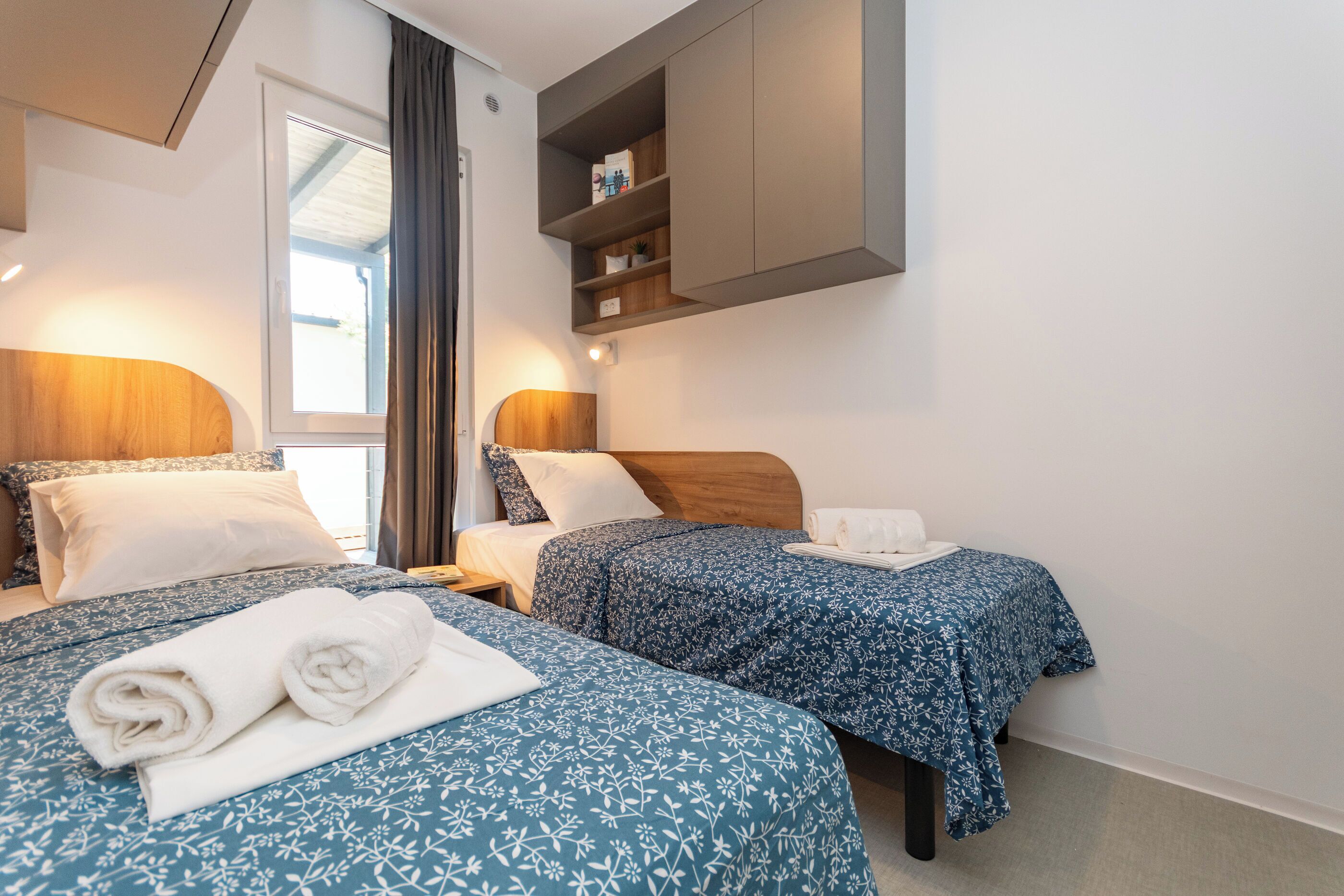 Hotellihuone, jossa on kaksi sänkyä, puinen pääty, sisäänrakennettu hylly ja ikkuna, jossa on verhot.