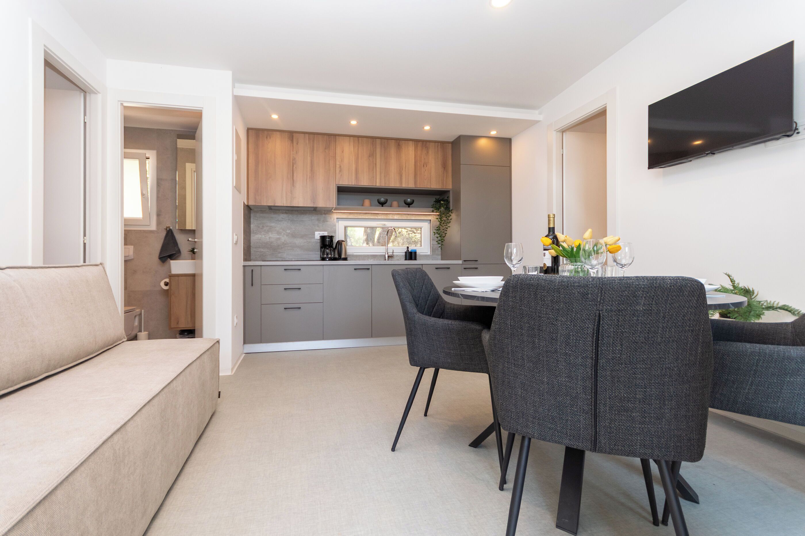 A modern kitchen with wooden cabinets, a dining area with grey upholstered chairs, and a flat-screen TV mounted on the wall.