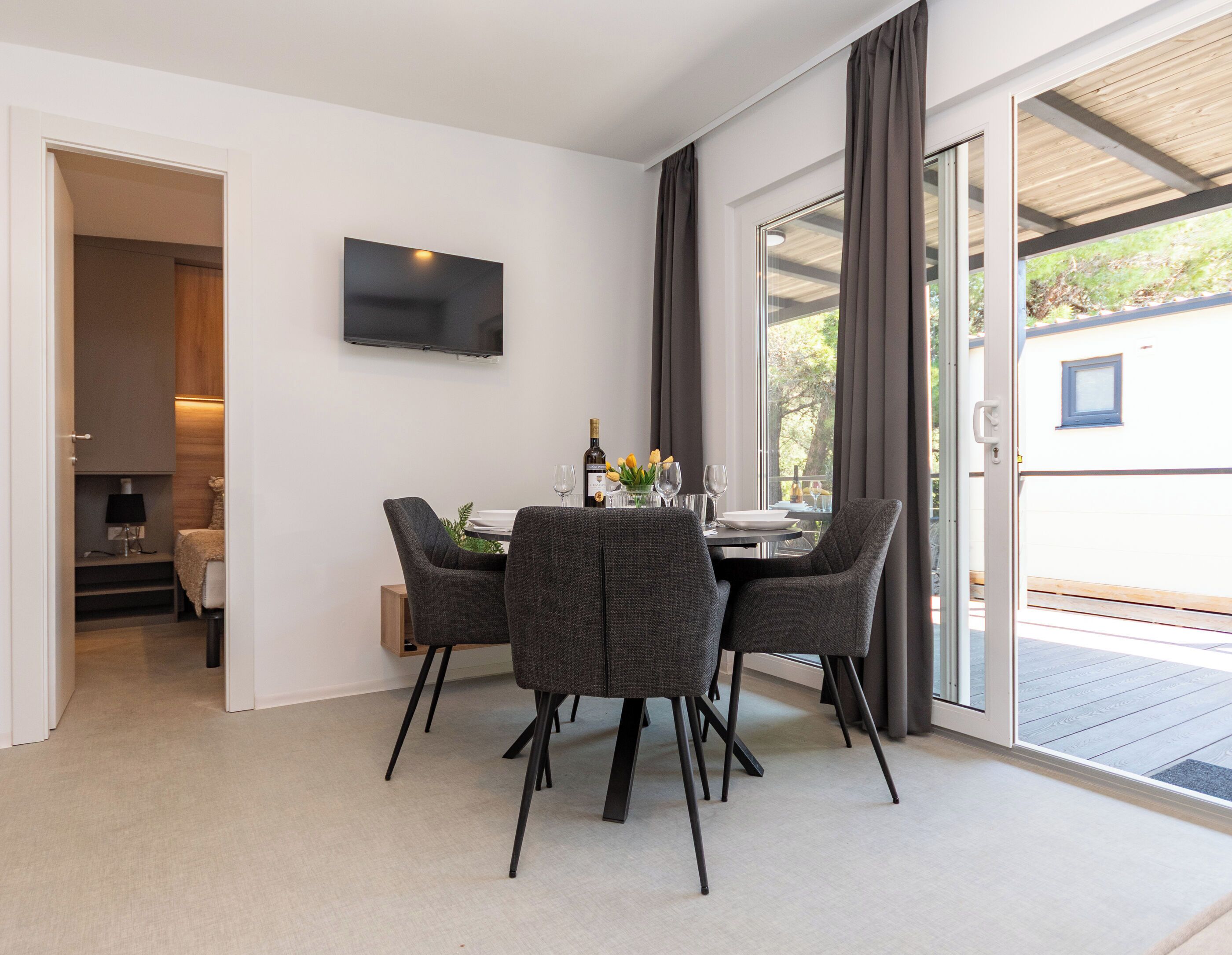 Un coin repas avec une table dressée pour quatre personnes, un téléviseur à écran plat fixé au mur et une porte-fenêtre coulissante donnant sur une terrasse extérieure.
