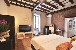Ein Hotelzimmer mit Bett, Fernseher, einer kleinen Küchenzeile, einem Bücherregal und einem Fenster mit Vorhängen.