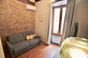 Ein Zimmer mit Backsteinwand, grauem Sofa, Fenster mit Vorhängen und Heizkörper.