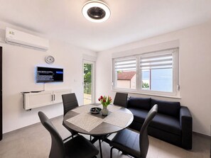 Ein modernes Wohnzimmer mit Essbereich, einem flachbildfernseher an der Wand, einer Couch und einem Fenster mit Jalousien.